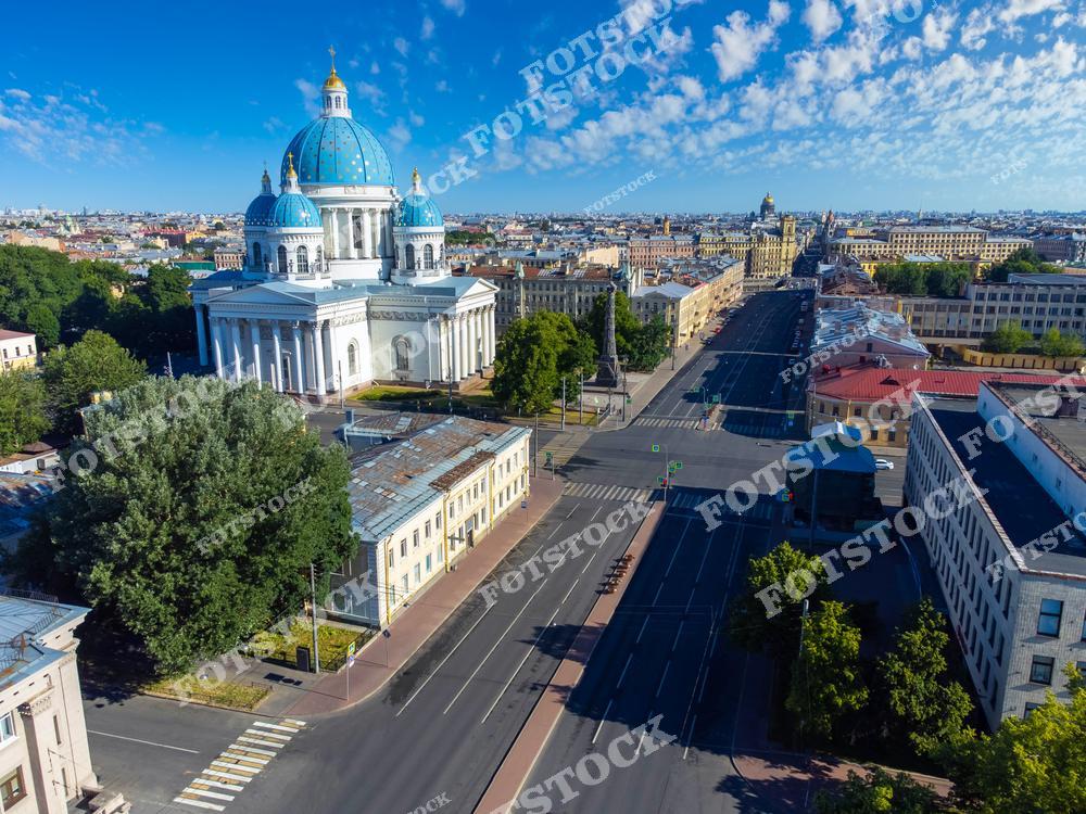 На фотографии изображен Троицкий собор с его характерными синими куполами, возвышающийся над Измайловским проспектом. Широкая улица, проходящая вдоль собора, открывает панораму города с его исторической архитектурой. Ясный летний день подчеркивает красоту фасадов и куполов. Собор, один из символов Санкт-Петербурга, является значимой достопримечательностью города.